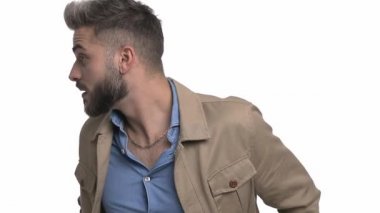 Kibirli havalı adam bej ceketini ayarlıyor ve etrafa bakıyor, bir davetiye yapıyor, parmaklarını yan yana gösteriyor, habersiz omuz silkiyor, el ele verip düşünüyor, stüdyoda beyaz arka planda tek başına duruyor.
