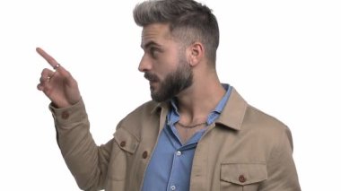 Kafası karışmış genç bir adam işaret ediyor ve yön veriyor, kafası karışık bir şekilde kaşınıyor, başını sallıyor, habersiz omuz silkiyor ve stüdyoda beyaz arka planda tek başına duruyor.
