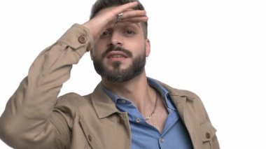 Bej ceketli, el ele tutuşmuş, elleri gözünün üstünde, arayarak, parmaklarını göstererek, gülüp, el sallayarak uzaktaki stüdyoda beyaz arka planda gezen tuhaf bir adam.