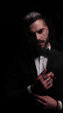 Siyah smokinli kendinden emin genç bir manken, şehvetli bir şekilde yan tarafa bakıyor ve poz veriyor, gizemli bir şekilde stüdyodaki siyah arka planda ayrılıyor.