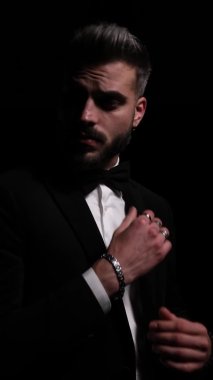 Zarif genç adamın dikey portresi siyah smokini ayarlıyor, boynu tutuyor ve yana bakıyor, gizemli bir şekilde stüdyodaki siyah arka planda ayrılıyor.