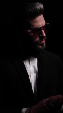 Siyah smokinli dramatik bir genç adam duygusal bir şekilde avuçlarını ovuyor ve yan tarafa bakıyor, güneş gözlüklerini tamir ediyor, parmaklarını gösterip siyah arka planda karanlıkta kayboluyor.