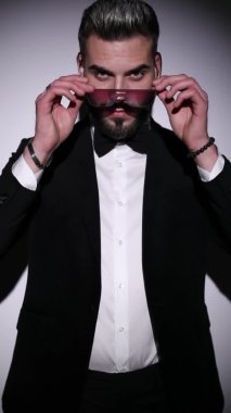 Seksi genç manken siyah kadife smokini ilikliyor, güneş gözlüğü takıyor, kollarını kavuşturuyor, parmaklarını şıklatıyor ve stüdyoda gri arkaplanı açıyor.
