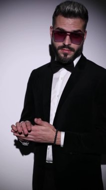 Kendine güvenen bir adamın dönüşünün dikey portresi, papyon takması, siyah kadife smokin giymesi, kollarını kavuşturması ve stüdyoda gri arka planda sessizlik hareketi yapması.