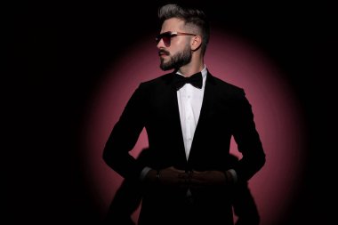 Yakışıklı moda işadamı ceketini kapatıyor, yana bakıyor ve pembe arka planda güneş gözlüğü takıyor.