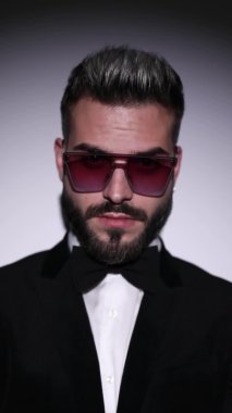 Güneş gözlüğü takan, el ele tutuşan, dudaklara dokunan, yan yana duran ve gri arka planda siyah kadife smokin giyen havalı genç adam.