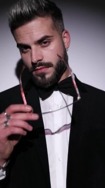 Yakışıklı, zarif bir adamın dikey portresi. Gözlük takıyor, yana bakıyor, siyah kadife smokin giyiyor ve gri arka planda şehvetli bir şekilde boynunu tutuyor.