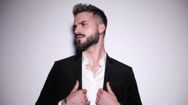 Yanlara bakan seksi genç manken, siyah kadife smokini ayarlıyor, el ele tutuşuyor, dudaklara dokunuyor ve stüdyoda gri arka planda poz veriyor.