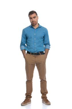 Tarafsız bir ruh hali içinde sıradan bir genç adam ellerini beyaz arka planda cebine sokuyor.