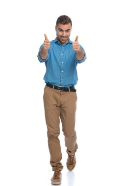 Genç, sıradan bir adam kameraya doğru yürüyor ve beyaz arka plana karşı coşkuyla baş parmak kaldırıyor.