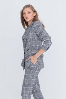 Gri kareli takım elbiseli şık bir modelin yan görüntüsü. Ceplerinde el ele tutuşmuş, stüdyoda açık gri arka planda poz veriyor ve yürüyor.