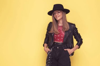 Genç ve güzel sıradan bir kız elleri ceplerinde poz veriyor ve sarı arka planda gülümsüyor.