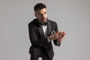 Siyah smokinli düşünceli genç bir adam dirseğini dizine dayamış, yana bakıyor ve stüdyoda açık gri arka planı düşünüyor.