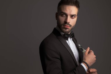 Siyah smokinli, papyonlu, mendilli, omuzlarının üstünden bakan ve stüdyoda koyu gri arka planda poz veren zarif bir genç.