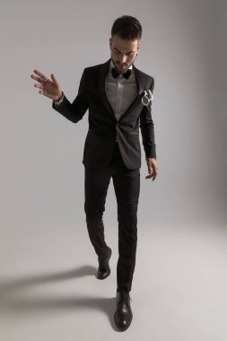 Siyah smokinli, papyonlu ve mendilli zarif bir genç stüdyoda açık gri arka planda yürürken aşağıya ve yana bakıyor.