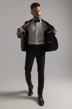 Ciddi bir genç adam siyah smokin giyiyor. Tarafını arıyor ve kaşlarını çatıyor. Stüdyoda açık gri arka planda yürüyor.