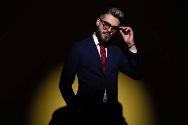 Donanma mavisi takım elbiseli, güneş gözlüğü takan, el ele tutuşan ve stüdyoda sarı arka planda moda ışığı altında poz veren havalı bir adam.
