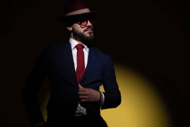 Sakallı, şapka takan, güneş gözlüklü şık bir adam. Takım elbisesinin kenarına bakıyor, sarı arka planda moda ışığı altında poz veriyor.