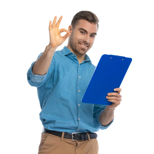 年轻英俊的休闲男子做一个确定的标志 并拿着一个蓝色的剪贴板与白色背景 — 图库照片
