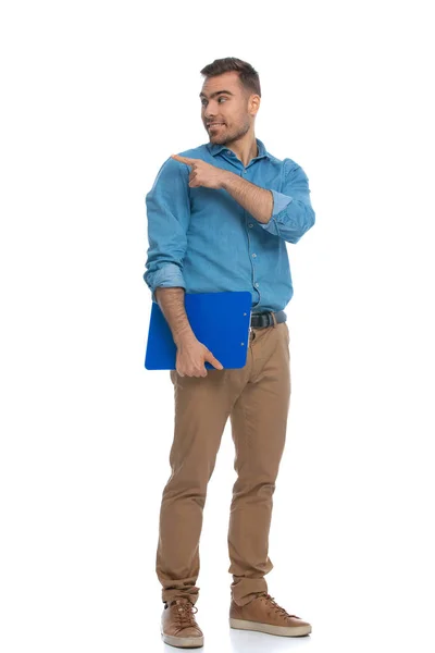 性感而随意的男人站在一边 指向相反的方向 拿着一块白色背景的蓝色剪贴板 — 图库照片