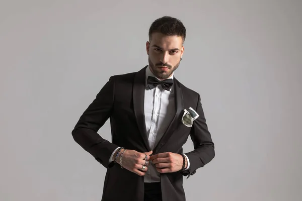 自信満々の男性モデルが蝶ネクタイとハンカチを身につけ黒タキシードを着けてスタジオで薄灰色の背景に官能的にポーズ — ストック写真