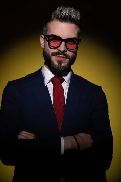 happy young businessman in navy blue suit crossing arms, posing and smiling, standing in a fashion light on yellow background in studio