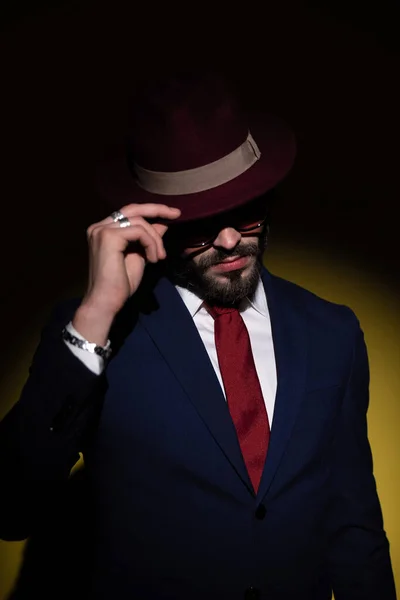 dramatic elegant man in navy blue suit pulling hat over face and hiding, looking down and posing in a fashion light on yellow background