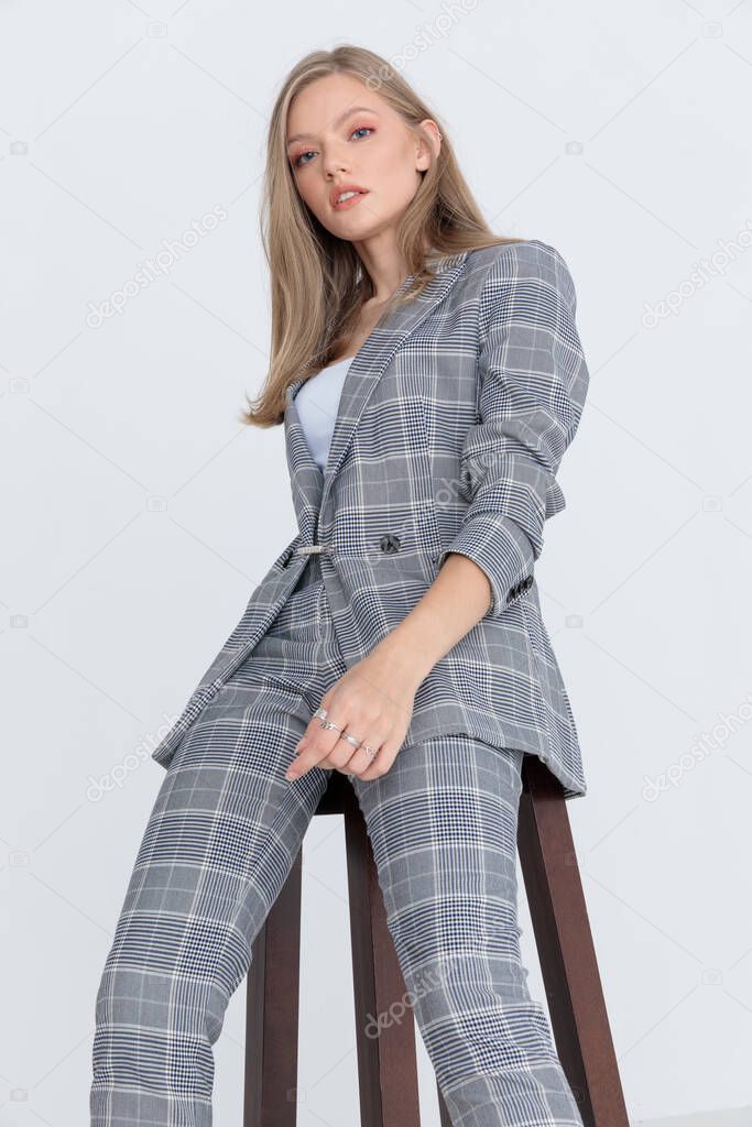 cool blonde model in gray checkered suit looking down and confidently posing, sitting on light gray background in studio