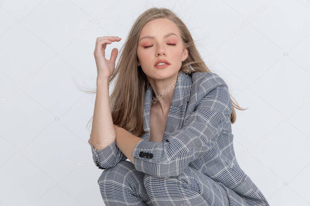 crouched young lady with closed eyes in gray checkered suit holding arms in a fashion pose on light gray background in studio