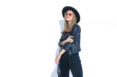 Beyaz arka plan, şapka ve güneş gözlüklü, kollarını tutup poz veren havalı bir genç kızı sunar.