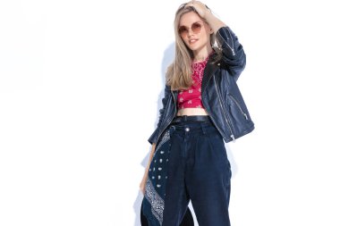 Deri ceketli sarışın kız saçlarını tarıyor, saçlarını tarıyor, gülümsüyor ve stüdyoda beyaz arka planda izole bir şekilde poz veriyor.