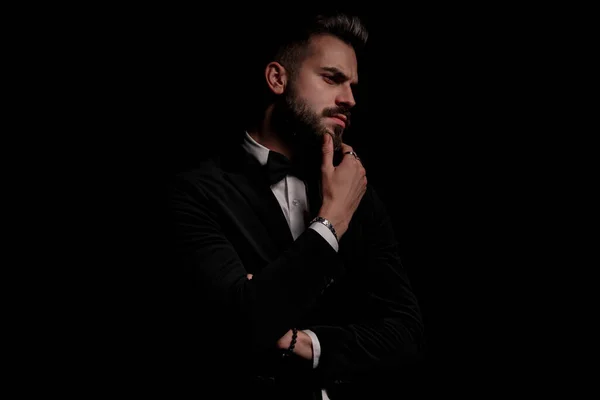 dramatic unshaved guy in black velvet tuxedo holding hand to chin and thinking, looking to side and posing on black background in studio