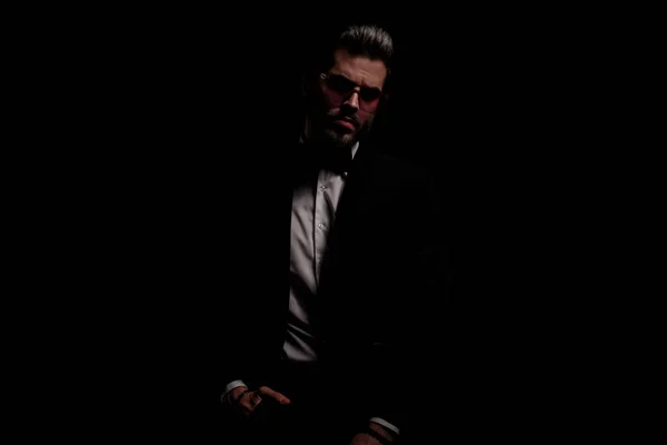 arrogant fashion guy in black velvet tuxedo holding hands in pockets and posing in a fashion light on black background in studio