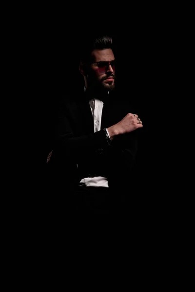 cool bearded guy in black velvet tuxedo with sunglasses posing on black background and looking to side