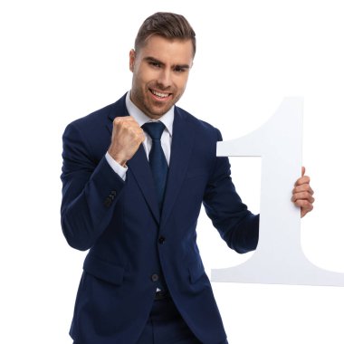 Hevesli genç model yumruğunu kaldırıp zaferi kutluyor, gülümsüyor ve stüdyoda beyaz arka planda bir numaralı tabelayı tutuyor.