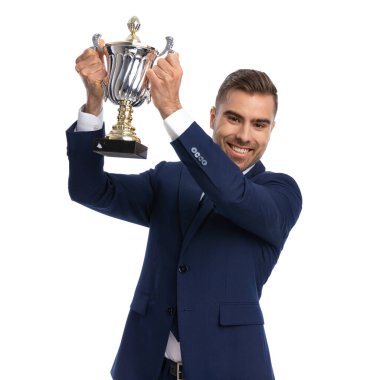 Lacivert takımlı gururlu genç manken elinde gümüş kupa tutuyor, gülümsüyor ve stüdyoda beyaz arka planda izole bir şekilde poz veriyor.