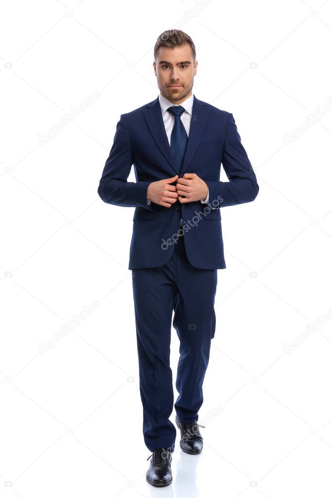attractive unshaved young model walking and arranging navy blue suit isolated on white background in studio