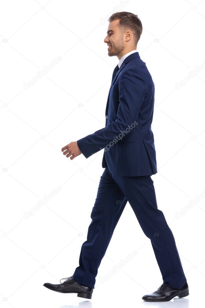 full body picture of elegant young man in navy blue suit looking to side and walking isolated on white background in studio