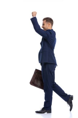 Heyecanlı mavi takım elbiseli genç işadamının yan görünüşü havada kol kola tutuşup zaferi kutluyor, stüdyoda beyaz arka planda tek başına yürüyor.