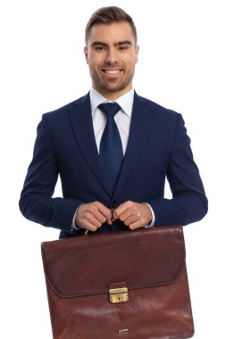 Zarif lacivert takımlı gülümseyen genç adamın portresi gururla bavul tutuyor ve stüdyoda beyaz arka planda izole bir şekilde poz veriyor.