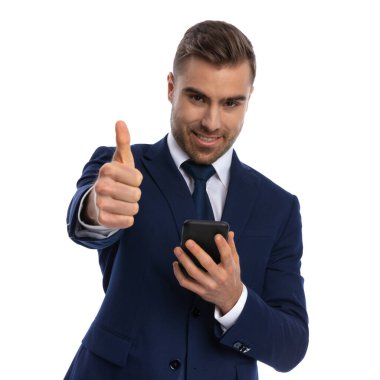 Lacivert takımlı yakışıklı sakallı adam beyaz arka planda duruyor, gülümsüyor, telefonu tutuyor ve stüdyoda baş parmak hareketi yapıyor.