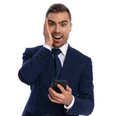 Heyecanlı mavi takım elbiseli manken harika haberler okuyor, el ele tutuşuyor ve suratını buruşturuyor, hevesle gülümsüyor ve stüdyoda beyaz arka planda izole bir şekilde poz veriyor.