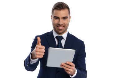 Lacivert takım elbiseli heyecanlı genç adam elinde sekme tutuyor ve başparmak işareti yapıyor, gülümsüyor ve stüdyoda beyaz arka planda izole bir şekilde poz veriyor.