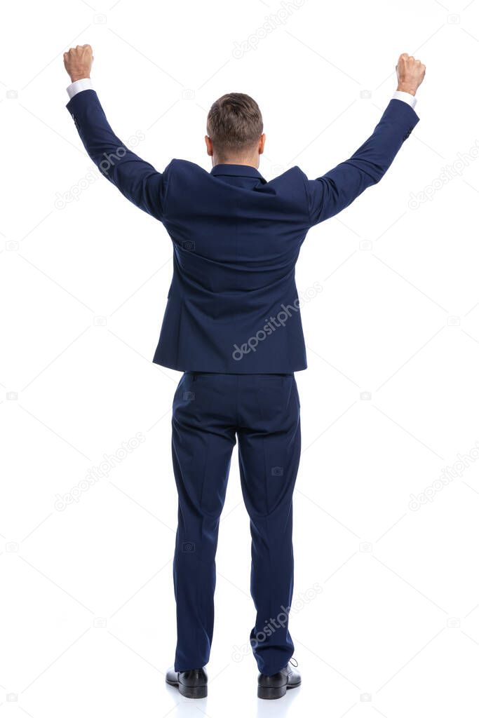 back view of elegant businessman in navy blue suit holding hands in the air and celebrating victory, standing isolated on white background in studio