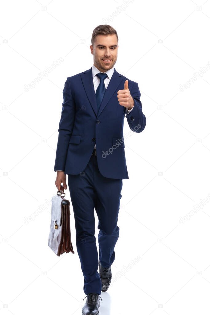 handsome young guy on white background wearing elegant navy blue suit, holding suitcase and making thumbs up gesture, walking isolated in studio