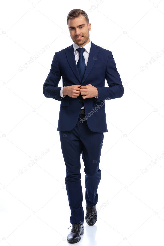 full body picture of unshaved young man smiling and adjusting navy blue suit, walking isolated on white background in studio