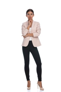 Pembe ceketli, kollarını kavuşturmuş, el ele tutuşup düşünen, ayakta duran ve stüdyoda beyaz arka planda poz veren düşünceli genç bir iş kadını.