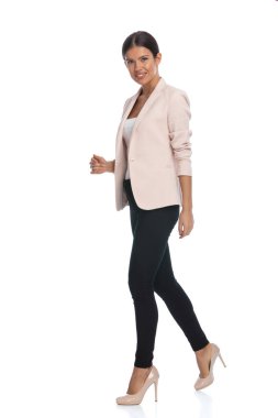 Pembe ceketli çekici esmer kızın yan görünüşü. Stüdyoda beyaz arka planda, tüm vücuduyla gülümseyen ve yürüyen.