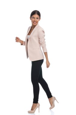 Pembe ceketli, gülümseyen ve omzunun üzerinden bakan güzel bir kadının yan görüntüsü. Stüdyoda beyaz arka planda tek başına yürüyor.