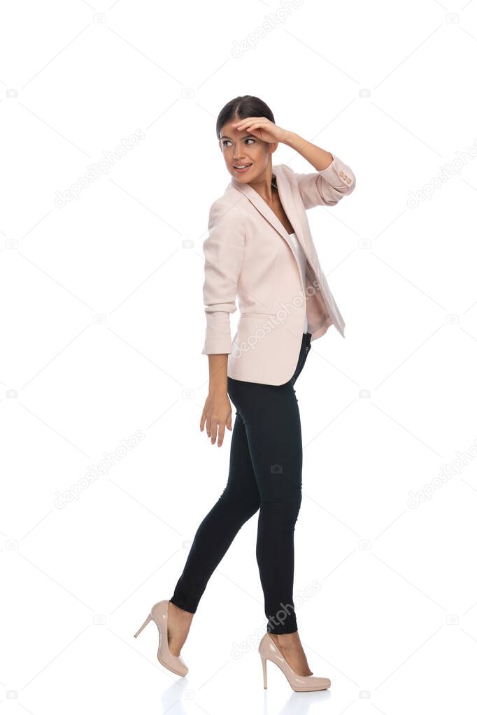 side view of smart casual girl in pink jacket holding hand to forehead, watching at a distance, military saluting and looking over shoulder, walking isolated on white background in studio